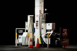 Gas station pumps are situated under dim lighting, with various hoses and digital displays visible. Orange safety cones are placed nearby, and signage advertises a Friday pie day deal.