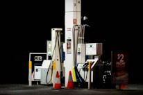 Gas station pumps are situated under dim lighting, with various hoses and digital displays visible. Orange safety cones are placed nearby, and signage advertises a Friday pie day deal.