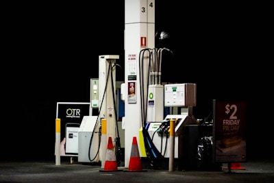Gas station pumps are situated under dim lighting, with various hoses and digital displays visible. Orange safety cones are placed nearby, and signage advertises a Friday pie day deal.