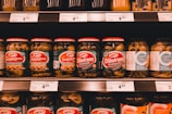 A supermarket shelf filled with various jars of pickles. The jars have red and white lids and green labels featuring brand names and pickle varieties such as Sandwich Savers and Crunch'ems. Price tags are visible on the shelf below, with some prices marked.