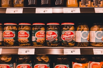 A supermarket shelf filled with various jars of pickles. The jars have red and white lids and green labels featuring brand names and pickle varieties such as Sandwich Savers and Crunch'ems. Price tags are visible on the shelf below, with some prices marked.