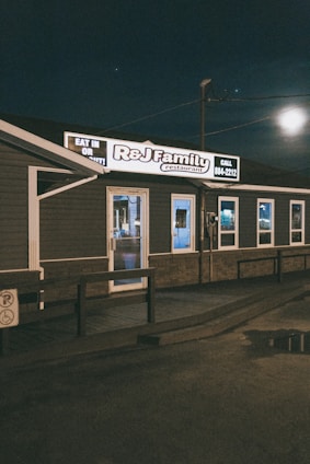 A small, family-owned restaurant with dark exterior walls and a well-lit sign displaying the name 'R&J Family Restaurant.' The building is set against a nighttime sky with some visible reflections on wet pavement.