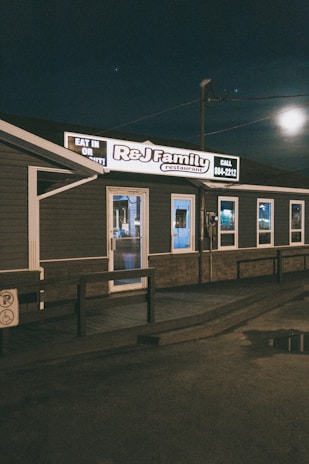 A small, family-owned restaurant with dark exterior walls and a well-lit sign displaying the name 'R&J Family Restaurant.' The building is set against a nighttime sky with some visible reflections on wet pavement.