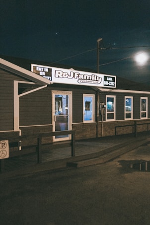 A small, family-owned restaurant with dark exterior walls and a well-lit sign displaying the name 'R&J Family Restaurant.' The building is set against a nighttime sky with some visible reflections on wet pavement.