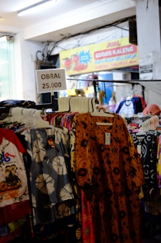 A variety of colorful clothing items hang on racks in a store. A sign above the racks reads 'OBRAL 25.000', indicating a sale. In the background, there's a banner advertising canned gudeg, a traditional dish from Jogja.