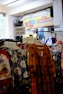 A variety of colorful clothing items hang on racks in a store. A sign above the racks reads 'OBRAL 25.000', indicating a sale. In the background, there's a banner advertising canned gudeg, a traditional dish from Jogja.