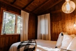 A cozy wooden cabin interior featuring a bed with white sheets and a black-and-white checkered blanket. The room is warmly lit with natural light coming through a window, alongside a glowing light bulb. White curtains with simple black patterns hang next to the window, and pillows are neatly arranged on the bed.