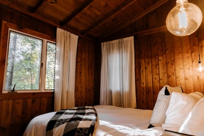 A cozy wooden cabin interior featuring a bed with white sheets and a black-and-white checkered blanket. The room is warmly lit with natural light coming through a window, alongside a glowing light bulb. White curtains with simple black patterns hang next to the window, and pillows are neatly arranged on the bed.