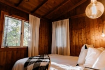 A cozy wooden cabin interior featuring a bed with white sheets and a black-and-white checkered blanket. The room is warmly lit with natural light coming through a window, alongside a glowing light bulb. White curtains with simple black patterns hang next to the window, and pillows are neatly arranged on the bed.