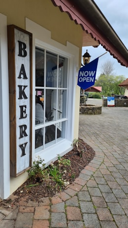 A quaint bakery with a sign reading 'BAKERY' next to a large window. A 'NOW OPEN' blue sign hangs near the entrance. The paved walkway curves gently, bordered by small plants and mulch. In the background, stone walls and a few trees can be seen under a cloudy sky.