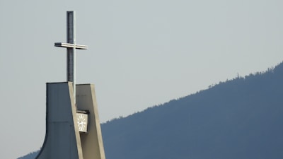 A tall structure with a cross on top, situated against the backdrop of a clear sky and a silhouette of a mountain range. The structure's angular design is modern, with a clock face visible below the cross.