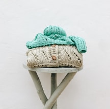 A knitted beige sweater with button details is neatly stacked, topped with mint green knitted fabric and a ball of yarn, on a round stool with crossed legs. The background is a plain white wall.
