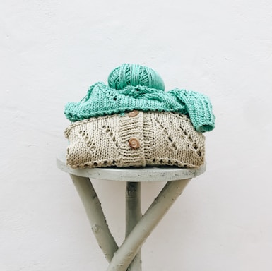 A knitted beige sweater with button details is neatly stacked, topped with mint green knitted fabric and a ball of yarn, on a round stool with crossed legs. The background is a plain white wall.
