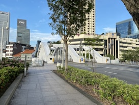 A modern cityscape featuring a white arched bridge spanning over a road. Surrounding the bridge are tall skyscrapers and office buildings with glass facades. There is greenery and flowering plants along the pathway leading up to the bridge. The sky is clear and blue, complementing the urban environment.