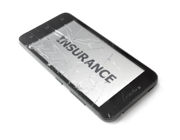 A smartphone with a severely cracked screen displays the word 'INSURANCE' on its screen. The device is black and appears to be placed on a white surface.