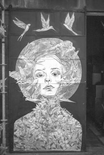 A monochromatic mural featuring a portrait of a woman's face intertwined with birds and floral patterns. The woman's expression is serene, and the birds are depicted flying through and around her, blending into her hair and body. The artwork is complex, with intricate lines and shading creating a sense of unity between the human and natural elements.
