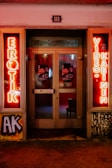 A storefront facade features illuminated neon signs with the words 'EROTIK' and 'VIDEO KABINEN'. The entrance consists of glass doors with posters displaying images and text. The surrounding area includes graffiti art on the walls, notably including the letters 'AK'. The scene is lit with a warm glow from the neon lights.