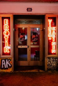 A storefront facade features illuminated neon signs with the words 'EROTIK' and 'VIDEO KABINEN'. The entrance consists of glass doors with posters displaying images and text. The surrounding area includes graffiti art on the walls, notably including the letters 'AK'. The scene is lit with a warm glow from the neon lights.