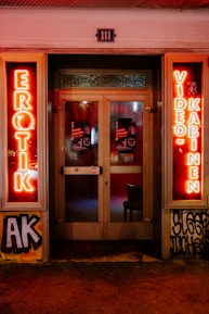 A storefront facade features illuminated neon signs with the words 'EROTIK' and 'VIDEO KABINEN'. The entrance consists of glass doors with posters displaying images and text. The surrounding area includes graffiti art on the walls, notably including the letters 'AK'. The scene is lit with a warm glow from the neon lights.