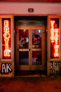 A storefront facade features illuminated neon signs with the words 'EROTIK' and 'VIDEO KABINEN'. The entrance consists of glass doors with posters displaying images and text. The surrounding area includes graffiti art on the walls, notably including the letters 'AK'. The scene is lit with a warm glow from the neon lights.