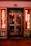 A storefront facade features illuminated neon signs with the words 'EROTIK' and 'VIDEO KABINEN'. The entrance consists of glass doors with posters displaying images and text. The surrounding area includes graffiti art on the walls, notably including the letters 'AK'. The scene is lit with a warm glow from the neon lights.