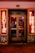 A storefront facade features illuminated neon signs with the words 'EROTIK' and 'VIDEO KABINEN'. The entrance consists of glass doors with posters displaying images and text. The surrounding area includes graffiti art on the walls, notably including the letters 'AK'. The scene is lit with a warm glow from the neon lights.