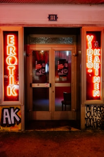 A storefront facade features illuminated neon signs with the words 'EROTIK' and 'VIDEO KABINEN'. The entrance consists of glass doors with posters displaying images and text. The surrounding area includes graffiti art on the walls, notably including the letters 'AK'. The scene is lit with a warm glow from the neon lights.