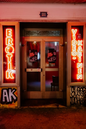 A storefront facade features illuminated neon signs with the words 'EROTIK' and 'VIDEO KABINEN'. The entrance consists of glass doors with posters displaying images and text. The surrounding area includes graffiti art on the walls, notably including the letters 'AK'. The scene is lit with a warm glow from the neon lights.
