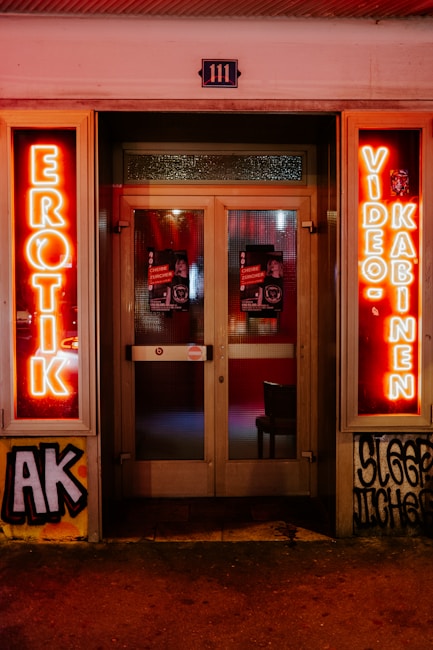 A storefront facade features illuminated neon signs with the words 'EROTIK' and 'VIDEO KABINEN'. The entrance consists of glass doors with posters displaying images and text. The surrounding area includes graffiti art on the walls, notably including the letters 'AK'. The scene is lit with a warm glow from the neon lights.