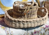 A woven basket with a handle is adorned with colorful beads and contains fabric with floral patterns. The basket rests on a table covered with embroidered cloth, featuring intricate floral designs.