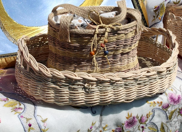 A woven basket with a handle is adorned with colorful beads and contains fabric with floral patterns. The basket rests on a table covered with embroidered cloth, featuring intricate floral designs.