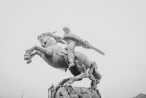 A monochrome statue of a man wielding a sword while riding a rearing horse, positioned against a foggy sky.