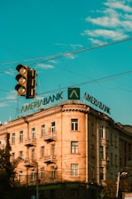 A multi-story building with a classic architectural style features a large, prominent sign on its roof displaying the word 'Ameriabank'. The sky is a vibrant blue with scattered clouds, and there is a traffic light in the foreground.