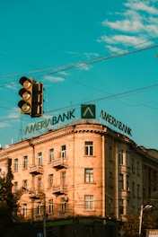A multi-story building with a classic architectural style features a large, prominent sign on its roof displaying the word 'Ameriabank'. The sky is a vibrant blue with scattered clouds, and there is a traffic light in the foreground.