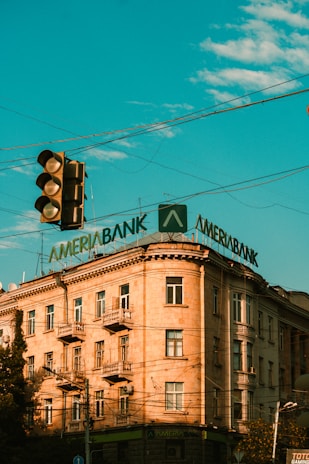 A multi-story building with a classic architectural style features a large, prominent sign on its roof displaying the word 'Ameriabank'. The sky is a vibrant blue with scattered clouds, and there is a traffic light in the foreground.