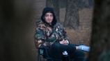 A person is sitting on a foldable chair in a wooded area. The individual is wearing a camouflage jacket with a hood pulled over their head. The background consists of several tree trunks, and the ground is covered in brown leaves or soil.