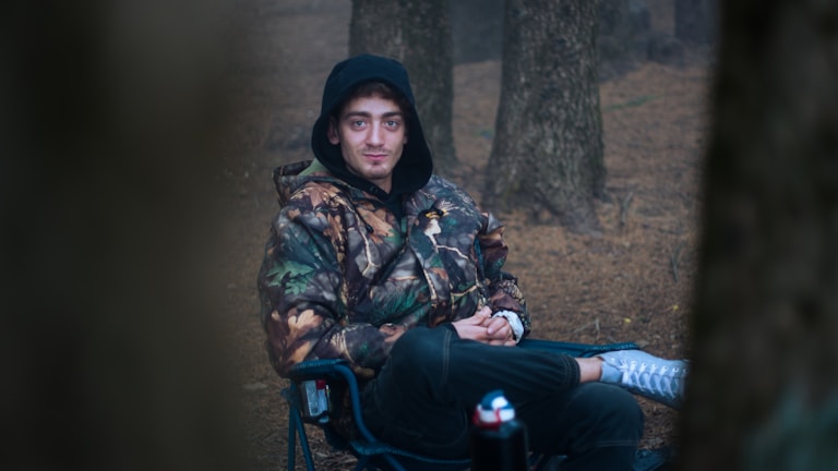 A person is sitting on a foldable chair in a wooded area. The individual is wearing a camouflage jacket with a hood pulled over their head. The background consists of several tree trunks, and the ground is covered in brown leaves or soil.