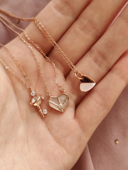 Three delicate gold necklaces rest on an open palm. The pendants feature intricate designs: one resembling a wand with a small crystal, another shaped like a geometric heart with inlaid mother-of-pearl, and the third a minimalist heart design with a pearl accent. The background is a soft, pink fabric with a smooth texture.