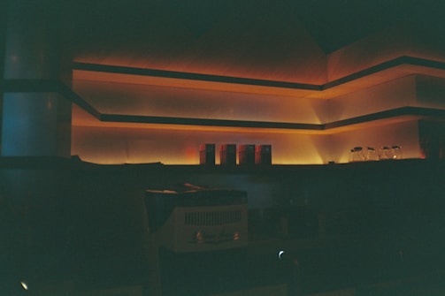 A dimly lit interior with warm, ambient lighting creating an inviting atmosphere. The shelves are illuminated with a soft orange glow, and several glasses and objects are arranged neatly. The lighting emphasizes the architectural lines and angles.