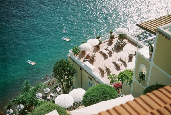 A coastal scene featuring a terrace overlooking the sea, with several white umbrellas and lounge chairs on the terrace. Below the terrace, there are people swimming in the clear, turquoise water. Green palm trees and well-maintained bushes add to the ambience of the location. The water sparkles under the sunlight.