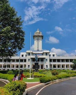 A grand, two-story white building with large windows and a prominent clock tower stands majestically against a vibrant blue sky. In front of the building, a well-maintained garden features lush greenery and a statue placed on a pedestal. Two individuals are walking along a path, one wearing a red outfit. The scene conveys a sense of tranquility and academia.