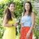 Two women standing in a garden setting, each holding a large wine glass. One woman is wearing a yellow dress, while the other is in a blue top with a red floral skirt. They are smiling and surrounded by lush green foliage.