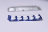 A close-up view of a blister pack containing a total of ten round, white pills or tablets. The blister pack is primarily blue, and it is placed on a flat, white surface. Another, partially blurred blister pack is visible in the background.
