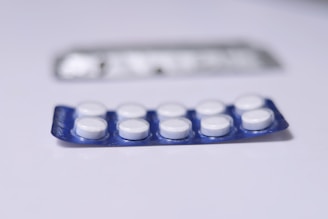 A close-up view of a blister pack containing a total of ten round, white pills or tablets. The blister pack is primarily blue, and it is placed on a flat, white surface. Another, partially blurred blister pack is visible in the background.