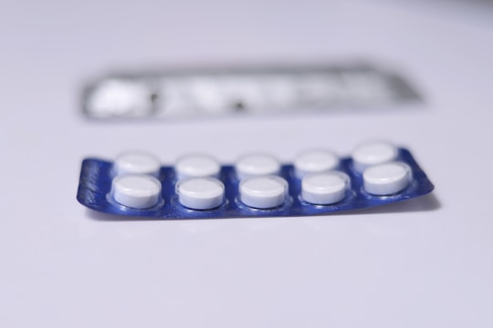 A close-up view of a blister pack containing a total of ten round, white pills or tablets. The blister pack is primarily blue, and it is placed on a flat, white surface. Another, partially blurred blister pack is visible in the background.