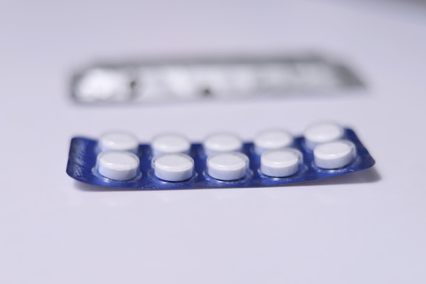 A close-up view of a blister pack containing a total of ten round, white pills or tablets. The blister pack is primarily blue, and it is placed on a flat, white surface. Another, partially blurred blister pack is visible in the background.