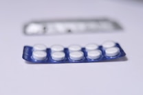 A close-up view of a blister pack containing a total of ten round, white pills or tablets. The blister pack is primarily blue, and it is placed on a flat, white surface. Another, partially blurred blister pack is visible in the background.