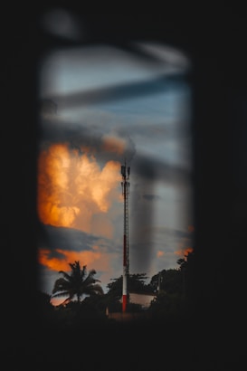 A telecommunications tower stands prominently against a backdrop of vibrant clouds at sunset. The scene is framed by silhouetted palm trees and darkened foliage, creating a contrast with the warm, glowing hues in the sky.