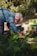 An elderly person with white hair and a striped shirt is bending down to tend to a raised bed garden filled with green leafy vegetables. The surrounding environment includes various gardening tools and lush greenery in the background.