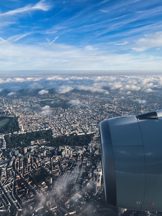 Aerial view of a cityscape partially covered by clouds, with a plane's engine visible in the foreground. The city is dense with buildings, and patches of greenery are scattered throughout. The sky above is bright with scattered clouds and a deep blue hue.